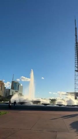 The video showcases a wide shot of a large, open public space in Brasília, Brazil, featuring a prominent telecommunications tower and a decorative fountain. In the foreground, a paved walkway with some greenery and benches creates a pedestrian area. The central focus is a multi-tiered fountain with several jets of water spraying upwards, creating a dynamic visual against the clear blue sky.

To the right of the fountain stands the tall, lattice-like telecommunications tower, a distinctive landmark of the city. A modern building is visible in the background to the left. The sun is low in the sky, casting long shadows and bathing the scene in a warm, late afternoon light, suggesting the time is around sunset. The sky is a vibrant blue with a few scattered white clouds.

Two silhouetted figures are walking along the walkway in the mid-ground, near the fountain, adding a sense of scale and human presence to the scene. The overall atmosphere is peaceful and serene, highlighting the architectural and natural beauty of the urban landscape. There is no visible text in the video.