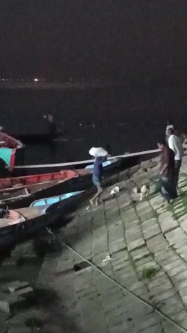The video shows a night scene on a waterfront with several small boats docked along a stone embankment. At least three people are visible, including one carrying a white sack on their head. Two of them are working on or near the boats. The boats appear to be made of wood and are painted in various colors including red, blue, and green. The water is dark, and there are faint lights visible in the distance across the water.