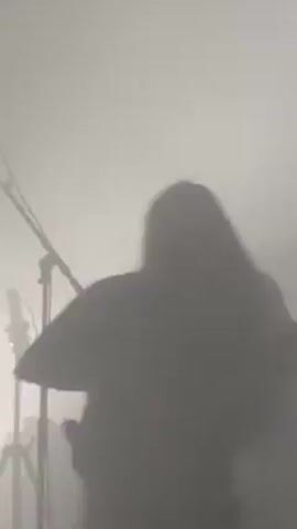 A silhouette of a long-haired person playing a guitar is visible through heavy smoke. The person is facing left and strumming the guitar. Another partial silhouette is on the right, and a microphone stand is visible to the left.  Bright stage lights are behind them.
