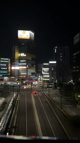 The video shows a nighttime view of a city street with buildings and traffic. In the foreground, a road runs horizontally across the frame, with cars driving on it. The buildings on either side of the road are illuminated with lights, creating a cityscape. The sky is dark, indicating that it is nighttime. A few cars are visible on the road, with their headlights and taillights on. Some of the buildings have large illuminated signs. The overall impression is that of a busy urban environment at night.