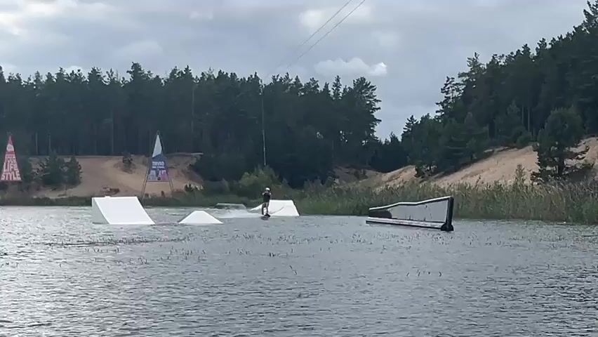 A wakeboarder is being pulled by a cable system across a body of water. The wakeboarder is wearing a helmet and a dark shirt. They are in the process of performing a trick, as evidenced by the spray of water around them. In the background, there are several ramps and obstacles placed in the water, indicating a wake park. Beyond the water, there is a line of trees and sand dunes. The sky is overcast.