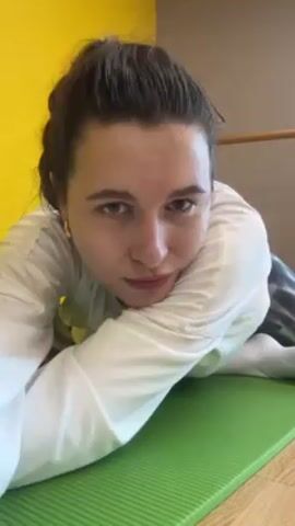 A person with fair skin and brown hair pulled back is lying on a green yoga mat on their stomach. They are wearing a white, long-sleeved shirt and tie-dye pants. The person props themselves up on their elbows and then rests their head in their hand. The background consists of a yellow and tan wall. A wooden ballet barre is visible behind the person.