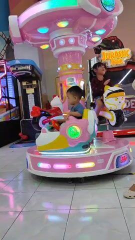 A low-angle, full shot video captured at eye level with two young children riding a pink, rotating amusement ride in an arcade. On the left, a toddler boy with short black hair, wearing a blue t-shirt, sits facing forward on a pink and teal carousel animal. He appears to be looking ahead. To his right and slightly behind, a young girl with long dark hair, wearing a maroon sweater, sits facing forward on a white and black carousel animal resembling a robot. She is looking to her left, her mouth slightly open. The carousel ride is adorned with colorful lights around its base and on its central column. In the background, several arcade games are visible, including one with a "Jurassic Park" theme on the right. The floor is tiled in a light gray and white checkered pattern. The overall scene depicts a playful and lively arcade environment.