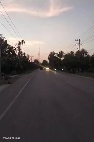 The video captures a first-person perspective of driving along a paved road at dusk in Putoh Sa, Indonesia. The sky displays soft hues of pale blue, pink, and orange with scattered clouds, indicating the early evening. A timestamp "2026.04.29 18:41" is visible in the bottom left corner, confirming the time.

On the left side of the road, utility poles with power lines run alongside a dense line of tropical vegetation, including numerous prominent palm trees. Further in the distance, a tall tower, possibly a cell tower, stands out against the horizon. On the right side, more trees and some distant structures are visible.

Several vehicles are seen driving in the opposite direction on the two-lane road. Their headlights are illuminated, piercing through the dimming light. At least two cars and one motorcycle are clearly identifiable among the approaching traffic. The scene suggests a tranquil evening drive or commute through a rural or semi-rural area.