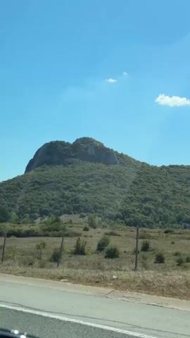 The video shows a landscape with a large, green, tree-covered mountain in the background. The sky is blue with a few scattered white clouds. In the foreground, there is a road with a metal guardrail. The camera appears to be moving, possibly from a vehicle. The overall scene is natural and undeveloped.