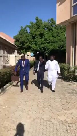 The video shows three men walking on a paved path outside. The man on the left wears a blue suit, a light-colored vest, a tie with a green pattern, and sunglasses. He carries a brown leather bag. The man in the middle wears a dark suit with a similar tie. The man on the right wears a long white shirt and pants. All three men are walking in the same direction. Behind them is a green tree, buildings, and a fence. The sky is blue.