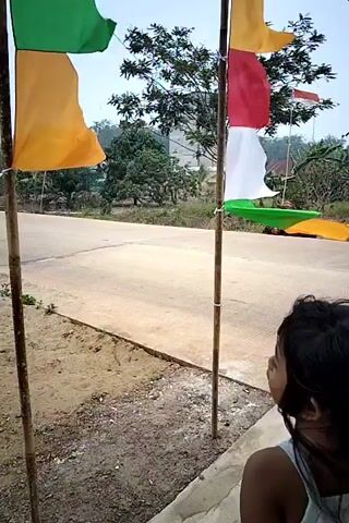A young girl with long dark hair, wearing a light blue tank top and dark shorts, stands on the left side of the frame, facing away from the camera. She is near a flagpole with Indonesian flags attached. In the background, a road runs horizontally, and on the right side of the road, a person wearing a headscarf rides a motorcycle with another person behind them. The environment appears to be rural, with trees and houses visible in the distance. The girl occasionally brings her hand to her mouth. The timestamp indicates the video starts at 00:00. The scene changes slightly to show a second person, a young female with dark hair pulled back in a bun, wearing a blue shirt, eating from a foil wrapper. Then, an older woman with graying hair, wearing a dark olive green t-shirt, stands facing right, looking away from the camera.