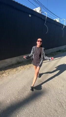 A woman with dark hair pulled back in a ponytail, wearing sunglasses, a black dress, a patterned jacket, and black sandals, walks on a gravel path. She is in motion, her arms are outstretched, and she appears to be talking or singing. The background consists of a dark, corrugated metal building and a clear blue sky. Sunlight casts shadows on the ground.