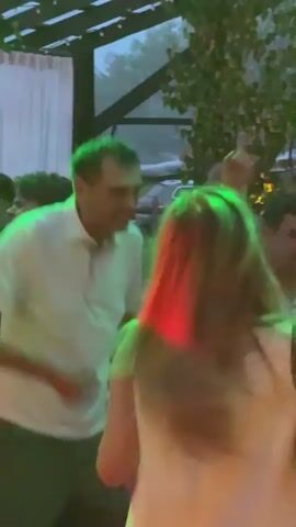 The video shows a group of people dancing outdoors at night. The lighting is low with red and green colored lights illuminating the scene. Trees and foliage are visible in the background. Several people are wearing white shirts. One person in the foreground has long brown hair and is wearing a white top. Another person in the background is wearing a white collared shirt and appears to be speaking. Some people are raising their hands in the air. The overall atmosphere appears to be festive.