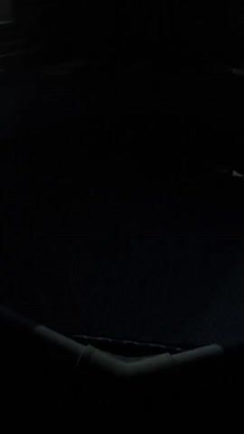 The video begins with a low-angle shot of a dark, enclosed space, possibly an outdoor area at night. A dark surface, possibly a trampoline, occupies the foreground, with a white corner support visible. The camera pans slowly to the left, revealing a dimly lit structure with a staircase leading up to an entrance. A light is visible from inside the building. The scene then shifts to show a dark wall with two windows. One window is brightly illuminated from within, while the other is darker. The camera continues to pan, showing more of the dark environment and the illuminated window. The video ends with a close-up of the illuminated window. The overall impression is one of a quiet, possibly deserted, outdoor space at night, with a single source of light emanating from a building.
