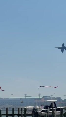 The video shows a gray and blue fighter jet in the sky. The jet ascends upward and banks slightly to the left. Below the jet are buildings and a dock with people standing on it. There are blue and white flags on the dock. A tree is visible in the right corner of the shot. The sky is a clear blue.