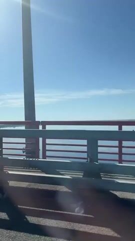 The video shows a view from a moving vehicle on a bridge. The bridge's railing is visible, featuring a combination of grey and red paint. The sky is bright blue with the sun shining, and a body of water is seen beyond the railing. The road surface is black and cast shadows.