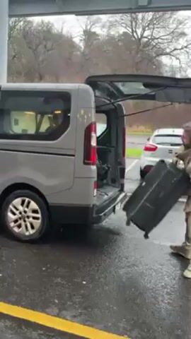 A man is shown loading a large black rectangular case into the open rear of a grey minivan. He's wearing beige pants, light tan sneakers and a tan coat with a fur lined hood. He has a backpack on as well. He struggles a bit to lift the heavy case and push it inside. The car is parked on a wet surface under a structure with green and white accents. There's a yellow painted line on the pavement next to the car. A silver car is parked to the right of the minivan. Trees can be seen in the background. It appears to be a cloudy, rainy day.