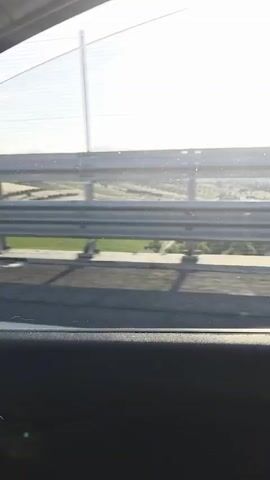 The video shows a view from inside a vehicle looking out at a sunlit landscape. A guardrail and a mesh fence are visible in the foreground, separating the vehicle from the exterior. Beyond the guardrail, rolling hills and fields can be seen under a bright sky. The sun is shining, creating a glare on the lens and obscuring some details of the distant scenery. The bottom portion of the video shows the interior of the vehicle, likely the car door panel. The footage appears to be a still shot or a very slow panning motion.