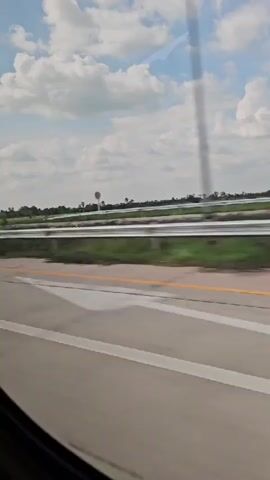 The video shows a view from a vehicle traveling on a highway. The road is paved with a yellow line on the right side and a white line on the left. Beyond the road, there is a green grassy median with a reflective barrier and then another lane of traffic. In the distance, there are trees and a few structures, including a water tower. The sky is blue with scattered white clouds. Several trucks are seen driving on the highway, including a white truck with a trailer, a blue and white truck with a long trailer, and a lighter-colored truck with a darker trailer. The camera is moving, giving a sense of motion.