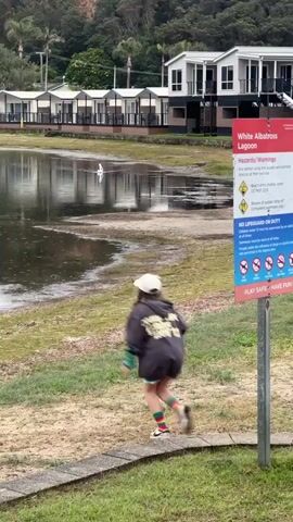 A young girl wearing a hat, a dark hooded sweatshirt with text, pink shorts, and striped socks is standing on a grassy bank. She is facing away from the camera and towards a body of water. In the water, a white bird, possibly a swan or pelican, is swimming. The bird is leaving a wake as it moves. In the background, there are several identical white buildings with dark trim and balconies, suggesting a resort or accommodation. A signpost with safety warnings is visible to the right of the girl. The environment appears to be a lagoon or calm body of water with muddy banks and surrounding vegetation.