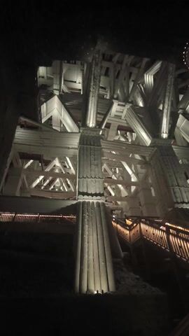 The camera pans upwards to reveal the intricate wooden structure of what appears to be an old mining tower. The structure consists of numerous interlocking beams and planks, all painted a light color. A wooden staircase with warm-toned lighting runs along the right side of the frame. Above the structure, two decorative wooden wheels with lights illuminating their rims hang from the ceiling. The overall environment is dimly lit and evokes a sense of age and industrial history.