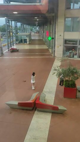 The video presents a static, overhead shot of a modern pedestrian walkway, likely in a commercial or public urban space in Prato, Italy. The ground is paved with reddish-brown tiles, featuring a prominent lighter-colored stripe running down the center.

In the foreground, a young person, dressed in a white short-sleeved dress and white sneakers, stands still in the middle of the walkway with their back to the camera. To the right, two modern benches with red seating surfaces and grey concrete bases are positioned. Further along the right side, near a building, a large red planter holds a green shrub, and two other individuals are partially visible standing nearby.

The walkway is flanked by multi-story buildings with contemporary architecture, characterized by large windows and glass facades. In the distance, on the left side of the walkway, a bright green cross sign, commonly used to denote a pharmacy, is clearly visible. Faint text, "Composizione," can be discerned on a building facade on the right. On the far left, a blurry orange sign shows partial text, possibly starting with "YAY...".

The scene appears to be set during the daytime, under clear weather conditions. The overall impression is one of a quiet, unhurried moment in a clean, modern urban environment. The audio includes spoken Italian, with a voice commenting, "Sì, sì, pena fuori dalla fotografia con la maglietta gialla" (Yes, yes, just outside the photo with the yellow t-shirt), suggesting an ongoing conversation or description of the surroundings.