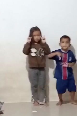 The video shows two young children standing in front of a plain white wall. The child on the left, a girl, is wearing a brown sweatshirt with a graphic print and gray pants. She has long dark hair and is making a peace sign with her hands near her head. The child on the right, a boy, is wearing a blue and red soccer jersey and shorts. He is standing with his arms bent at the elbows and his hands positioned as if ready to move. Both children appear to be looking at the camera. The floor in front of them is light-colored. There is a shadow cast on the wall behind the children. The video is short and appears to be a recording of the children posing for a picture or dancing.