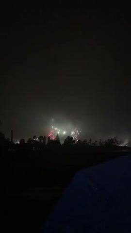 The video shows a nighttime scene with fireworks exploding in the distance over a silhouette of trees and other indistinct structures. The fireworks display is spread across the horizon, with bursts of light in white, pink, and green colors. There is a smoky or hazy atmosphere that diffuses the light from the fireworks. In the foreground, a dark area, possibly a field or lawn, is visible, and the corner of a dark blue umbrella with water droplets on it is present in the bottom right of the frame. The overall impression is of an outdoor event, likely a celebration, viewed from a distance.