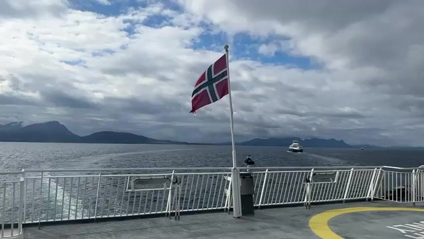 This media file captures a scenic view from the deck of a ferry as it travels through a body of water, likely a fjord, near Lødingen, Norway. The foreground is dominated by the ship's deck, featuring a white railing and a Norwegian flag fluttering on a flagpole. The flag, predominantly red with a blue cross outlined in white, is a prominent subject. Beyond the railing, the vast, grey-blue water stretches towards a distant, hazy shoreline.

The backdrop is a dramatic skyscape of heavy, grey clouds interspersed with patches of bright blue sky, suggesting a dynamic and possibly cool weather day. The mountainous terrain of the coastline is visible in the distance, appearing muted by the atmospheric conditions. A white ferry or ship is seen moving away from the viewer in the middle ground, leaving a visible wake on the water.

The scene conveys a sense of journey and perhaps contemplation, with the vastness of the sea and sky creating a tranquil yet imposing atmosphere. The time of day appears to be daytime, given the illumination from the sky. There is no visible text in the media file. The overall impression is one of a serene, perhaps slightly overcast, maritime journey in a beautiful, rugged landscape.