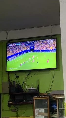 The video shows a television screen displaying a soccer match. The screen is mounted on a green wall. Below the television, there are several electronic devices and cables. In the foreground, a shelf holds some of these devices. To the right of the shelf, there is a glass-fronted cabinet with some items inside. The soccer match on the screen shows players in red and yellow uniforms on a green field, with spectators in the stands. The scoreboard indicates the time and score of the game.