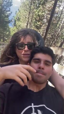 A video captures a woman and a man on a roller coaster. The woman is behind the man, her arms around him. The man is looking towards the camera with his eyes closed and a slight smile. The woman is wearing sunglasses and has curly brown hair. She is touching the man's face. The background shows trees and part of the roller coaster track. The video appears to be taken during the daytime, with natural lighting.