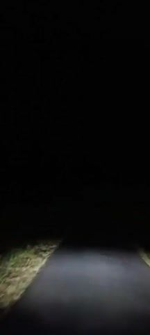 The video shows a dark, rural road at night. The headlights of a vehicle illuminate a small portion of the asphalt road and the grassy verges on either side. In the distance, there are a few scattered lights, possibly from houses or streetlights. The camera is mounted on a moving vehicle, as evidenced by the slight blur and the perspective of the road ahead. The surrounding environment appears to be undeveloped, with no significant structures or other vehicles visible. The overall impression is one of a quiet, isolated drive through the countryside after dark.