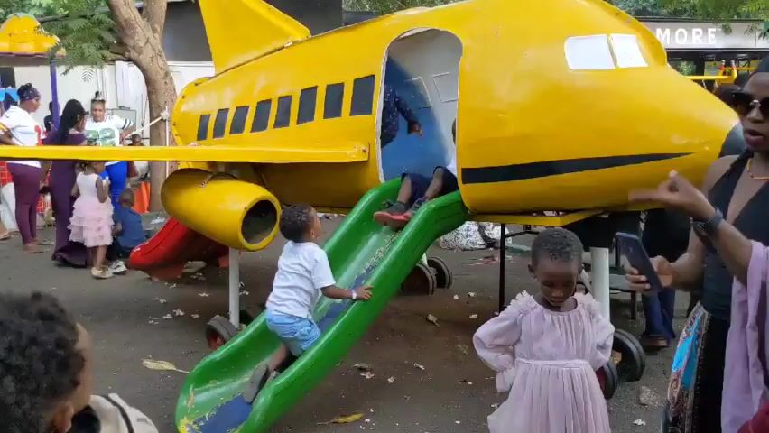 The video shows children playing on and around a large yellow airplane-shaped playground structure. The airplane has a slide attached to it, and several children are either climbing up the slide, sliding down it, or inside the airplane. A woman in sunglasses is recording the children with her phone. Other adults and children are visible in the background. The playground is located outdoors, possibly in a park, with trees and other playground equipment visible. The ground appears to be covered with dirt or gravel. Loud background noise is present, including music and perhaps other children playing.