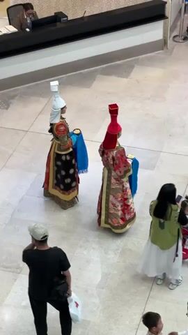 The video shows two individuals wearing traditional Mongolian attire with tall, ornate hats. One person wears a white hat and a black and gold robe, while the other wears a red hat and a red and gold robe with intricate patterns. Both are holding blue cloths. They are standing on a tiled floor, likely in a public indoor space such as an airport or a lobby, as suggested by a counter with a person seated behind it in the background. Several other individuals are visible, including a person with their back to the camera wearing a black shirt and a light-colored baseball cap, and another person wearing a green vest over a white dress. The scene appears to be a cultural display or performance.
