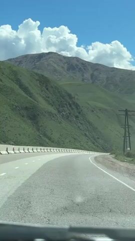 The video, shot from inside a moving vehicle, shows a road winding through a mountainous landscape on a sunny day. The asphalt road is lined with white concrete barriers on one side. Green, grassy hills rise steeply on both sides, leading up to a larger, rocky mountain in the background. White, fluffy clouds dot the bright blue sky. In the distance, a semi-truck with a blue trailer approaches on the road. Telephone poles stand beside the road, and some low-lying vegetation grows on the shoulder.