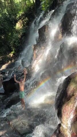 A person in green swim trunks stands on wet rocks at the base of a waterfall. They are reaching their arms up towards the cascading water, which creates a rainbow effect in the mist. Other people are visible in the background, further away from the waterfall. The environment is lush and green with trees surrounding the waterfall.