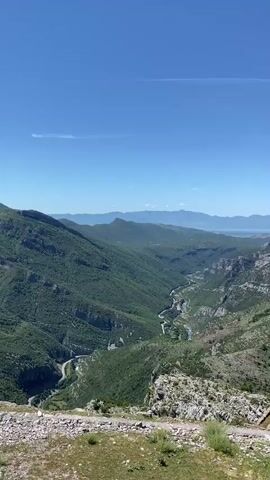 A vertical video shows a sweeping panorama of a vast, green, mountainous landscape under a clear blue sky. A winding river or road snakes through the valley, flanked by steep, tree-covered slopes. In the foreground, a rocky, grassy embankment suggests a viewpoint. The distant horizon is marked by a range of hazy mountains and a body of water, possibly a lake or the sea. The camera appears to be stationary, capturing a static, natural scene without any human activity or discernible objects other than the landscape itself. The timecode indicates the video is approximately four seconds long.