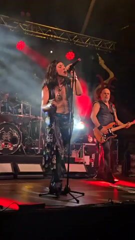 A male singer with long dark hair is on stage singing into a microphone. He is wearing a sleeveless vest, a necklace, and pants with skulls on them. To his right, a bald man plays a guitar. Behind them, a drum set is visible. The stage is lit with red and white lights.