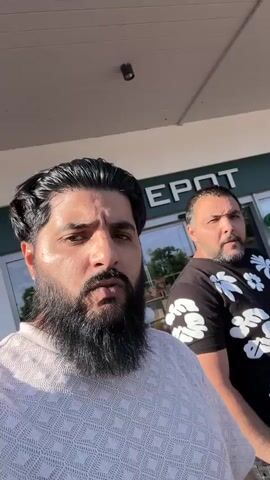 Two men are standing outdoors in front of a white building with large glass windows and doors. The man in the foreground on the left has a dark beard and hair, wearing a light-colored patterned shirt, and looks directly at the camera. The man on the right has shorter dark hair and a beard, wearing a black t-shirt with white floral patterns, and is looking to his left. A green sign above the building's entrance reads "DEPOT". A red sign partially visible on a glass window reads "40% AUF FAST ALLES". The man on the left gestures with his right thumb towards the building at the end of the video.