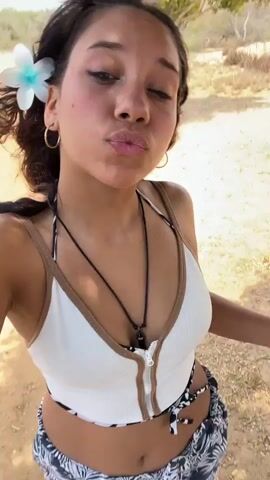 A young woman stands barefoot on a sandy or dirt surface, taking a selfie. She is wearing a white tank top, a patterned skirt, a necklace, and a flower in her hair. She has dark curly hair and is wearing hoop earrings. In the video she initially looks into the camera and then rotates the camera, putting her hand up to block the sun. The background appears to be outdoors, possibly a beach or park, with some vegetation visible.