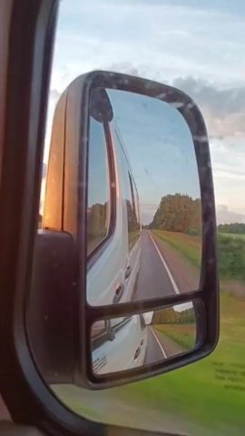 This video shows a reflection in a side-view mirror of a white van traveling on a rural road in Belarus. The scene captured in the mirror is of the road stretching into the distance, bordered by green fields and a line of trees. The sky is cloudy, and the lighting suggests it is either early morning or late afternoon. The time of day is consistent with the golden light reflecting on the van's body. There is no visible text in the video. The activity is travel, specifically driving on a road through a rural landscape. The overall mood is calm and peaceful, evoking a sense of journey and open road.