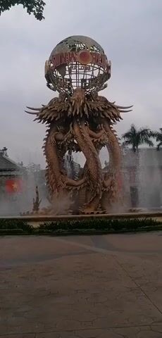 A static shot shows a large fountain sculpture in the foreground. The fountain is made of golden-colored dragons intertwined, supporting a metal globe with the words "SUNWORLD HALONG" on it. Water is spraying from various points of the sculpture, creating a mist around the base. The fountain sits on a brick-paved ground. In the background, there are buildings with traditional Vietnamese architectural elements, and a Vietnamese flag is visible on one of them. Palm trees are scattered around, and the sky is overcast. No people are immediately visible.