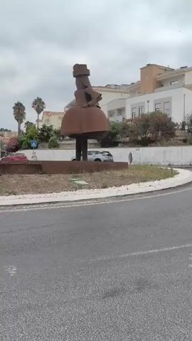 A rusty metal sculpture of a person wearing a hat and a large skirt stands in the center of a roundabout. Cars drive past the sculpture on a cloudy day. Buildings are visible in the background.