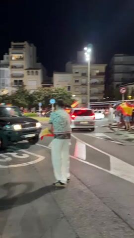 A black Hummer SUV is driving down a street at night. Spanish flags are attached to the vehicle and are being held by people on the sidewalk. The driver and a passenger can be seen inside the SUV. A person wearing a white shirt and white pants is standing next to the SUV, holding a Spanish flag. Another person in a blue shirt is leaning on the SUV. Buildings with lights on are visible in the background, suggesting an urban or town setting. The street appears to be paved and has lane markings. The lighting suggests it is nighttime.