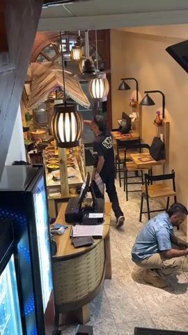 The video shows the interior of what appears to be a small restaurant or café, viewed from an elevated position, likely the top of a staircase. A man wearing a blue patterned shirt is crouched near a wall, working on electrical wiring, possibly with pliers. Another man in a black shirt stands near a counter, looking up, possibly observing something above.  The counter is made of light wood and has a woven texture on the front. It holds a computer and other items. A glass-fronted refrigerator stands to the left of the counter. Behind the counter is a buffet-style display with food items.  The restaurant has small tables and chairs, with decorative lamps above them. A wooden staircase with a decorative railing leads up to the second floor, which has a wood-paneled ceiling with recessed lighting and ceiling fans.  The floor near the man working on the wiring is tiled.