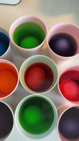 Here's a detailed description of the video:

The video shows a close-up, static shot of a collection of white paper cups arranged in a circular pattern on a white countertop. Each cup contains a different colored liquid dye and a hard-boiled egg submerged within it. The colors present include blue, green, purple, orange, red, and black.

A hand wearing a black glove appears and uses a silver spoon to lift one of the eggs from the dye. The egg appears to be in the process of changing color.