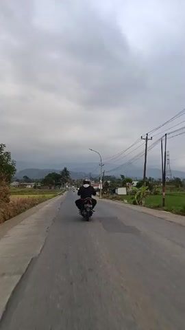 The video shows a person riding a motorcycle down a paved road. The person is wearing a helmet and a backpack. The road is lined with fields of crops on the left and some buildings and trees on the right. In the distance, there are mountains. The sky is overcast with clouds. Another motorcycle is visible further down the road. The footage appears to be shot from another motorcycle following behind.