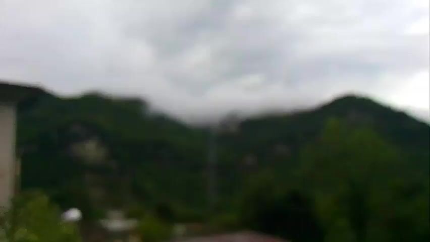 The video begins with a blurry shot of a mountainous landscape with green trees and cloudy skies. As the video progresses, the image sharpens, revealing more details. In the foreground, there is a portion of a building on the left and a few houses with reddish roofs. In the background, a tall transmission tower stands prominently between two densely forested green mountains. The mountains are covered in lush vegetation, and the cloudy sky suggests it might be overcast or raining. The overall scene depicts a rural or mountainous area, possibly a village nestled among the hills. There are no people visible in the video.