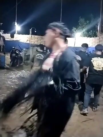 The video captures a nighttime outdoor event taking place in a muddy area, likely in Jenggrik, Indonesia. Artificial lights illuminate the scene, casting bright spots and shadows.

Numerous people, mostly men, are present and actively moving through the wet, muddy ground. Many individuals are dressed in black shirts, some featuring yellow graphic designs or logos on the back, though the text is not clearly legible. One man in a black shirt has a distinctive braided hairstyle.

At approximately the 0:03 mark, a person is seen falling or rolling in the mud. Other participants are observing or engaging in similar actions, suggesting a cultural performance or communal activity where interacting with the mud is part of the event.

In the background, a white building and temporary wooden fences or structures are visible. Some people are holding sticks or staffs. The ground's muddy condition indicates either recent rain or a deliberate setup for the event. The atmosphere appears to be lively and communal.