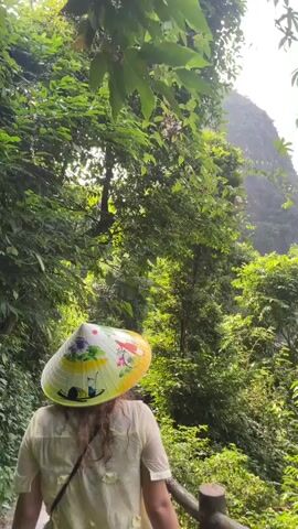 A person wearing a conical straw hat walks along a wooden walkway. The hat is decorated with colorful paintings, including a scene of boats. The person is wearing a light-colored shirt. The walkway is surrounded by lush green trees and foliage. In the background, there are large, steep, tree-covered karst mountains rising from the water. Several tour boats are visible in the water near the mountains. The sky is clear and blue. The camera pans to the right, revealing more of the seascape and the karst formations.