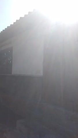 The video pans across the exterior of a building. The building appears to be made of concrete and wood, with a corrugated metal roof. There are two large wooden doors on the front of the building, and a small mailbox attached to the wall next to the doors. The building is situated in a grassy area, with trees visible in the background. The sun is shining brightly, casting long shadows. The video is filmed in a handheld style, and the camera is somewhat shaky.