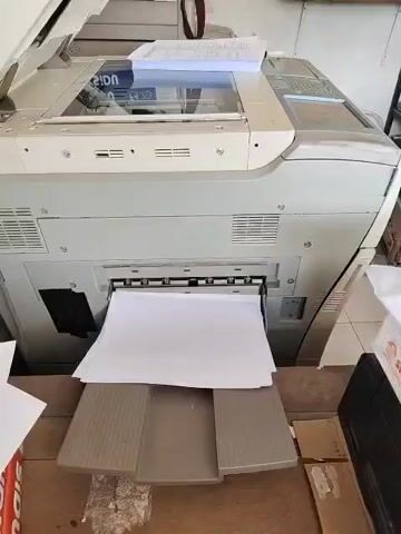 The video shows a close-up of a photocopier from a low angle, focusing on the output tray where multiple sheets of white paper are being ejected. The photocopier itself is large and appears to be an older model, with a beige and grey exterior. A stack of papers is visible in the document feeder on top of the machine. The output tray is partially filled with freshly printed pages. The surrounding environment appears to be an office or a copy shop, with hints of other office equipment and furniture visible in the background. The lighting is bright, suggesting an indoor setting. The action is continuous printing, with the paper being fed out from the machine.