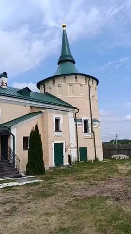 A camera pans slowly to the right, revealing a tall, cylindrical tower with a green conical roof and a gold cross at its peak. The tower is made of light-colored brick and features several arched windows and two green doors at its base. Adjacent to the tower is a beige building with a green roof and more windows. A tall, slender evergreen tree stands in front of the beige building. The ground in front of the structures is covered with dry grass. In the background, a wooden fence and utility poles are visible under a partly cloudy sky. The overall scene suggests an outdoor view of religious or historical architecture.