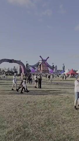 The video captures a lively outdoor event, likely a music festival, at Neustadt-Glewe, Germany. The central focal point is a large, elaborately designed stage structure resembling a giant windmill, adorned with vibrant purple lights on its blades. The structure also features the text "AIRBEAT ONE" prominently displayed on its facade.

Numerous people are visible in the foreground and background, milling about in a festive atmosphere. They appear to be enjoying the event, with some individuals dancing and others observing the surroundings. The crowd is diverse, suggesting a public gathering.

The scene is set during the daytime, under a clear, bright sky, indicating fair weather. The ground appears to be a grassy field, characteristic of an outdoor festival site. Smoke or fog effects are present around the stage, adding to the visual spectacle. The overall mood conveyed is one of excitement and celebration.